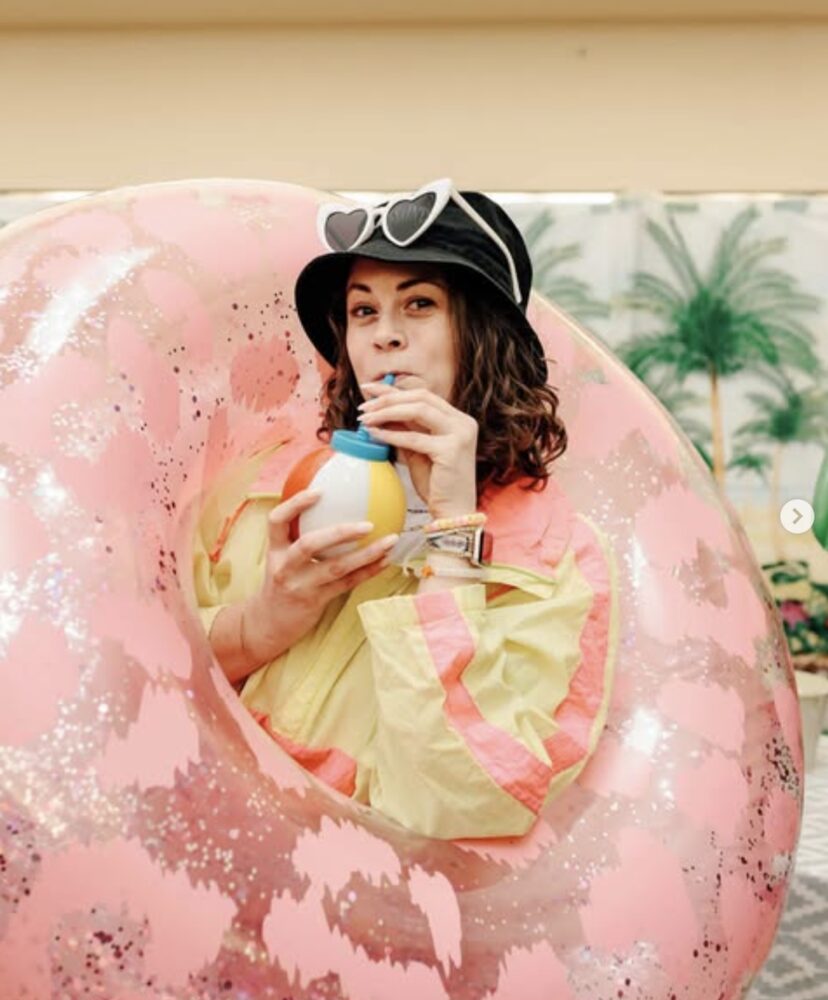 Leah in a giant pink pool float, bucket hat and heart sunglasses, sipping from a beach-ball cup